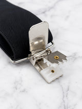 A close-up of a black woven suspenders with a heavy duty silver metal clip and white plastic release mechanism, resting on a white marble surface.