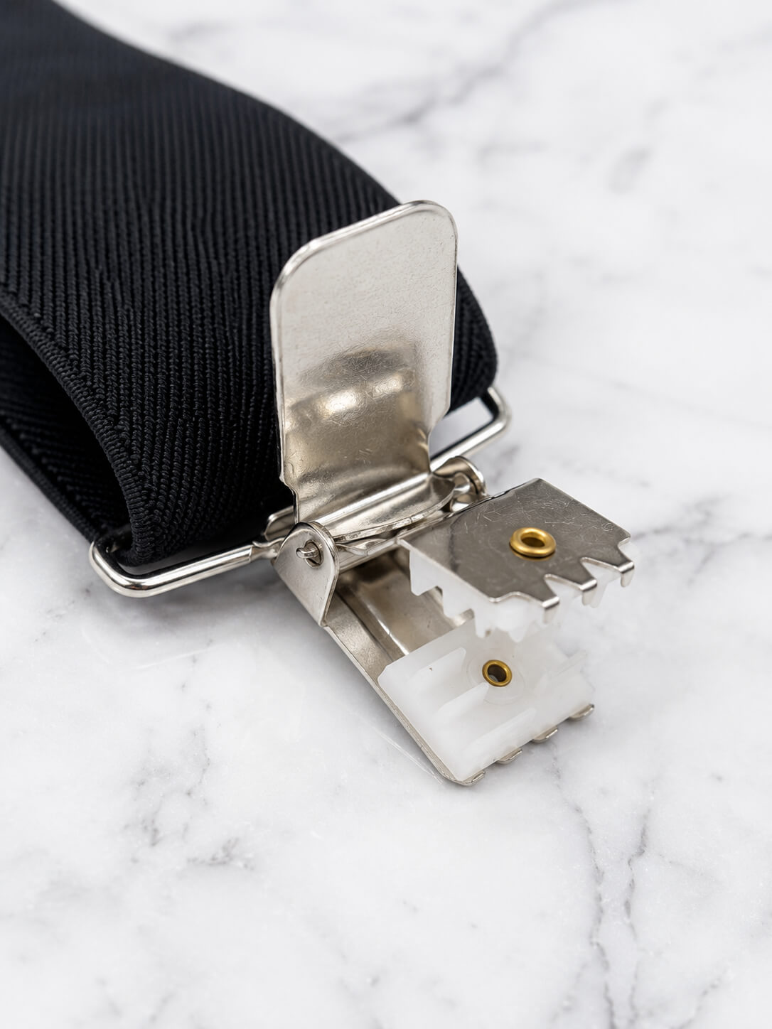 A close-up of a black woven suspenders with a heavy duty silver metal clip and white plastic release mechanism, resting on a white marble surface.