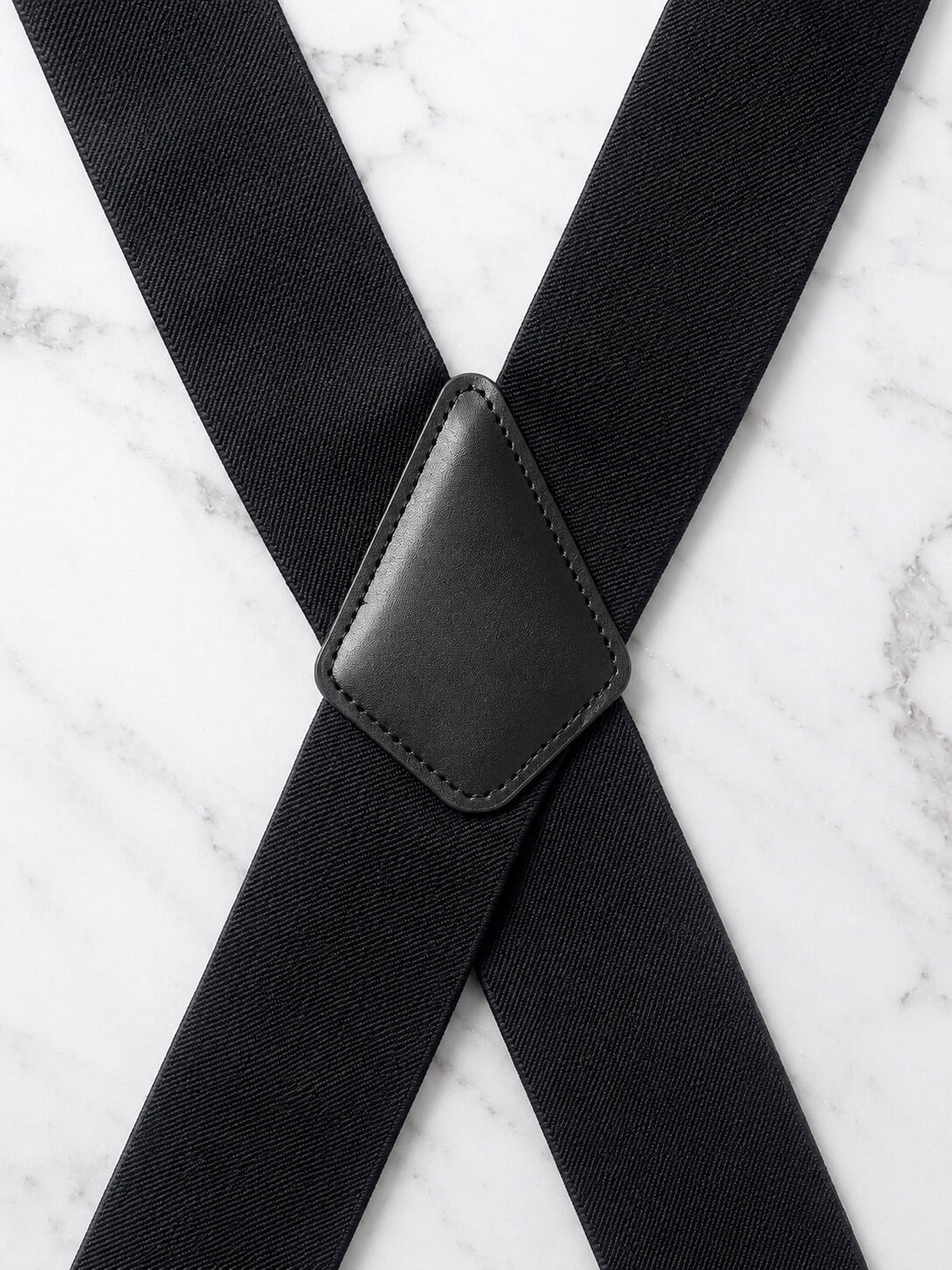Black suspenders with a textured fabric strap and a stitched black leather-like central connector, laid out on a white marble surface.
