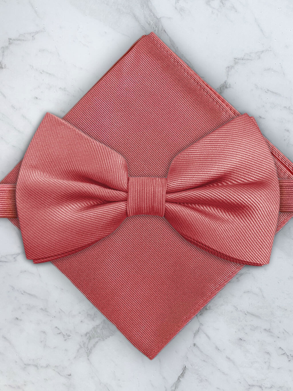 A coral-colored silk twill bow tie with a pocket square, displayed on a white marble surface.